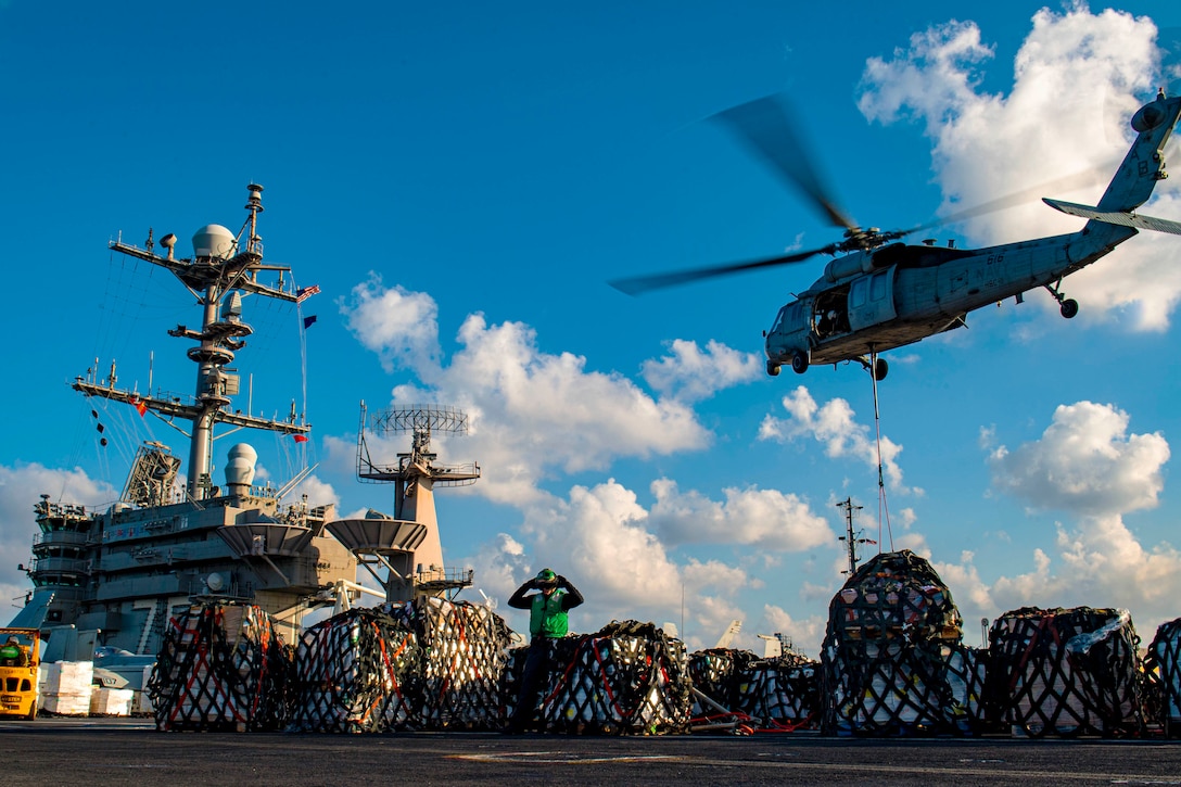 A helicopter with a pallet of supplies attached hovers over the deck of a ship.