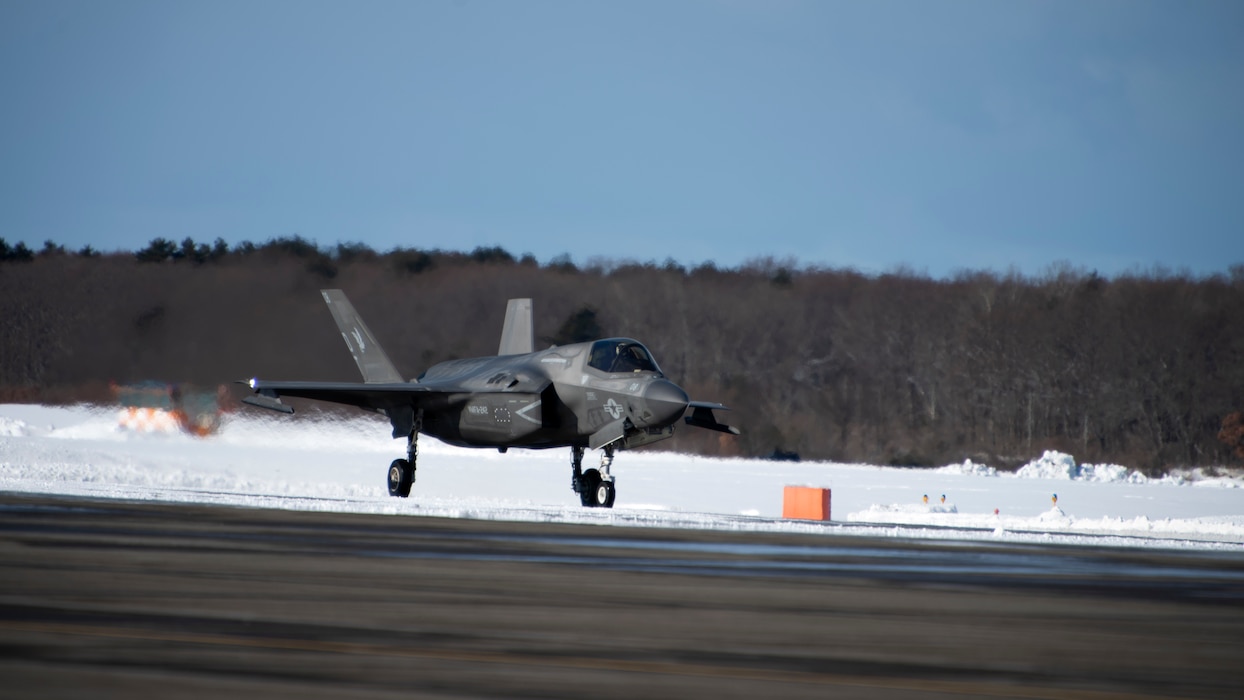 A grey aircraft drives across a flightline with a snowy landscape behind it