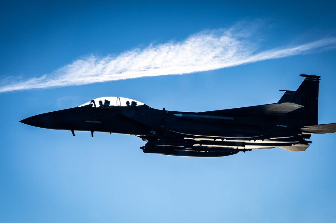 An aircraft flies in blue sky under a linear cloud.