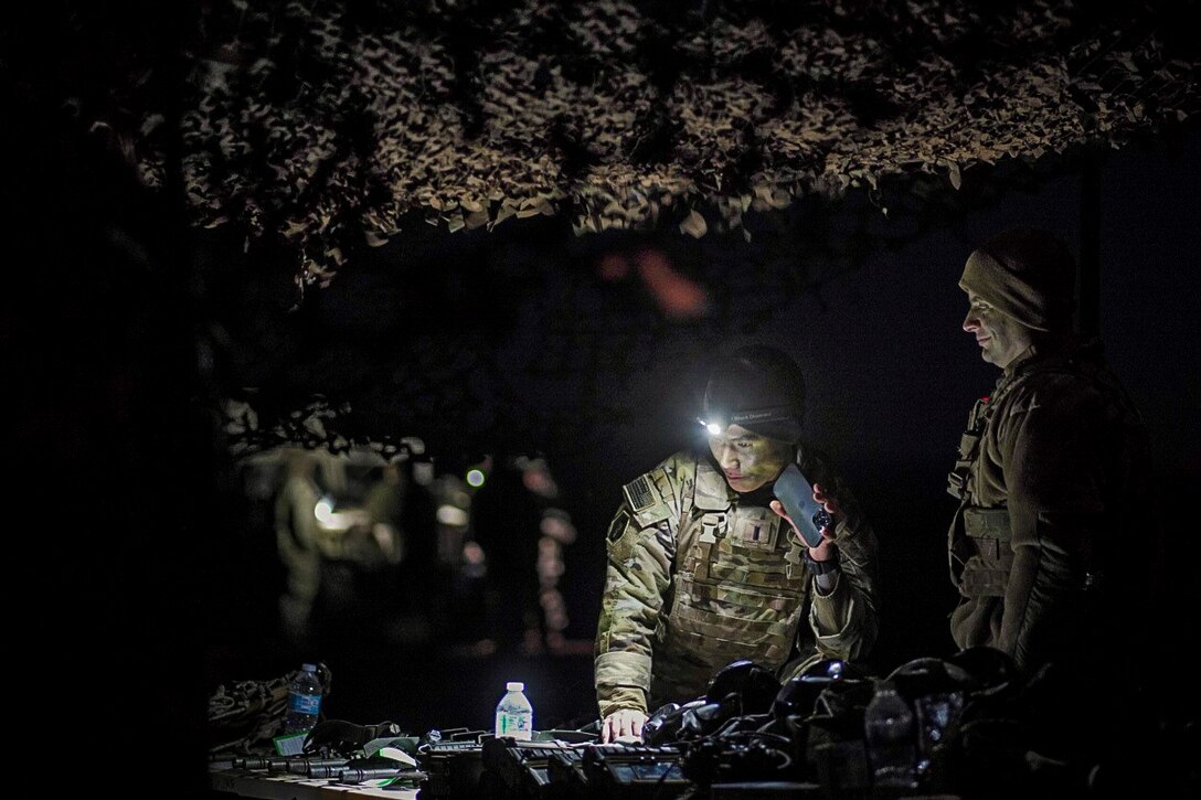 A soldier wearing a helmet with a flashlight leans on table while a fellow soldier stands next to him in the dark.