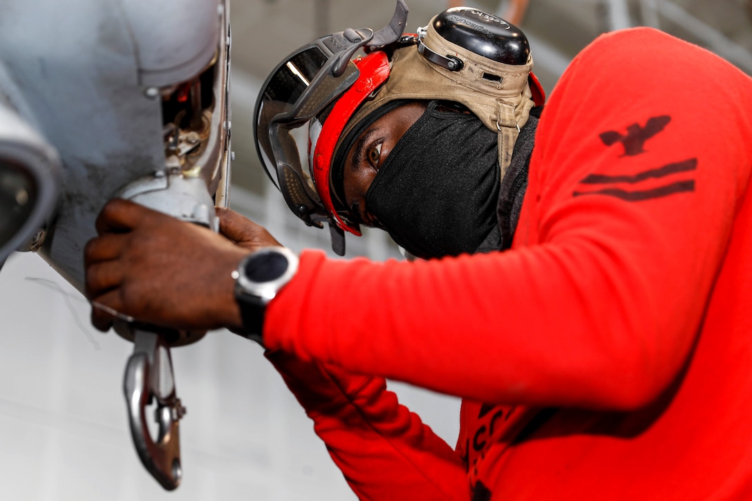 A sailor wearing a face mask and helmet conducts maintenance on a helicopter aboard a ship at sea.