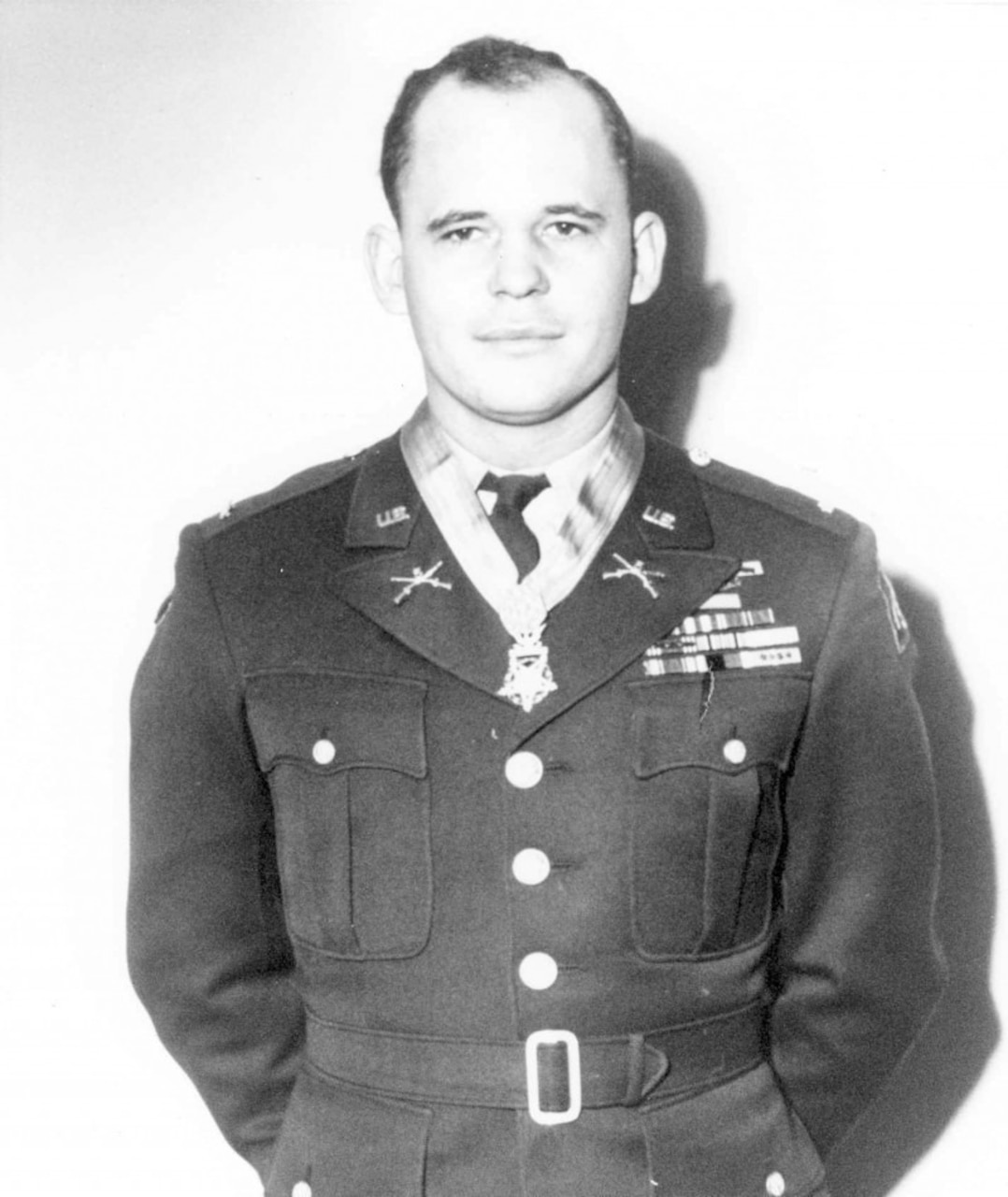 A soldier in dress uniform and medal around his neck stands with his hands behind his back.