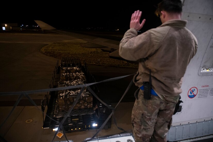 A uniformed service member directs the movements of a utility vehicle which carries cargo pallets.