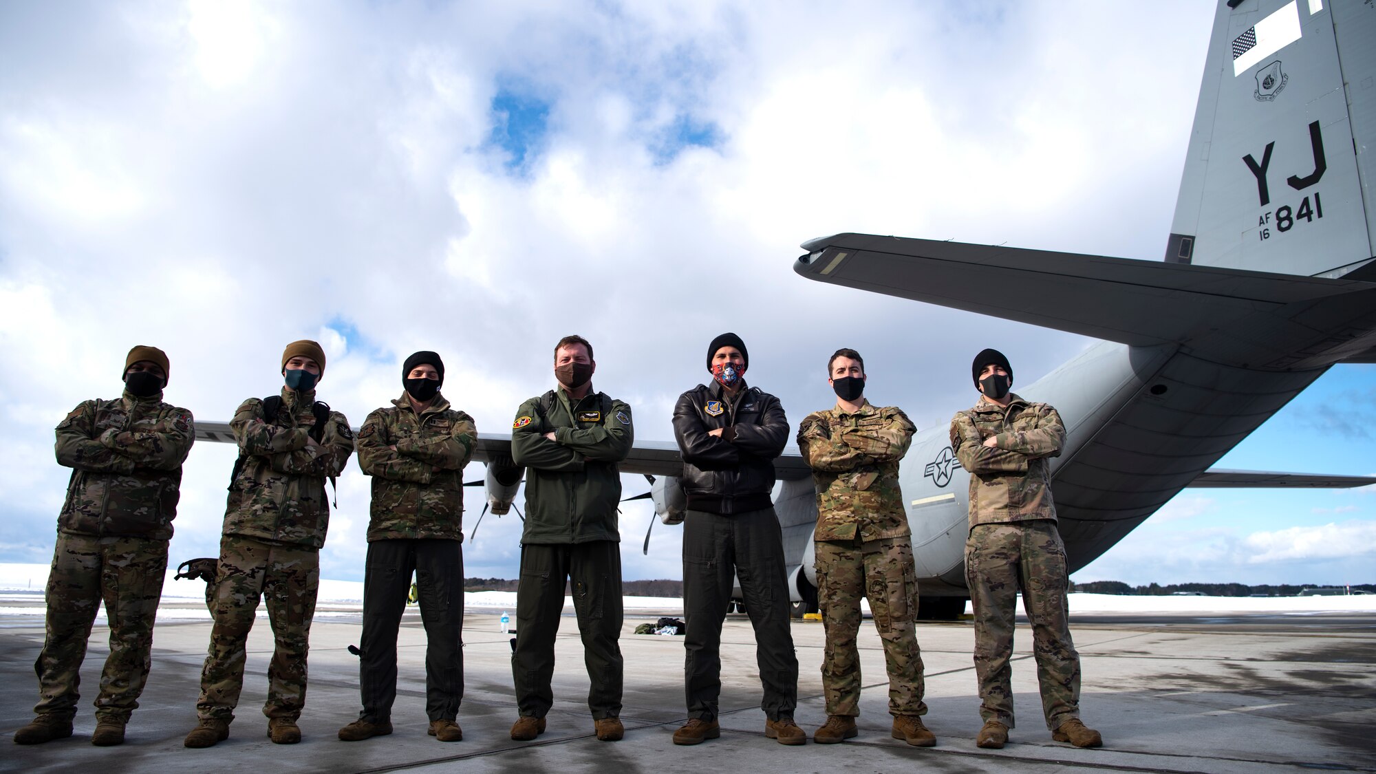 6 men in flight suits (green and OCP) stand to the side of a large aircraft with a mountain in the background on a snowy day