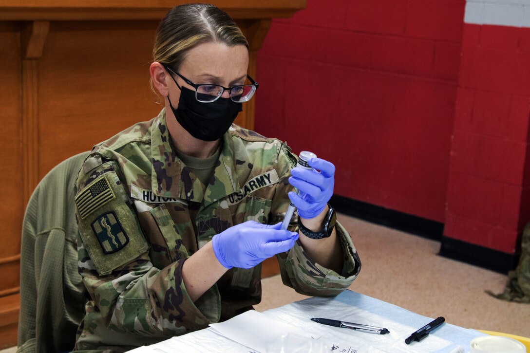 A soldier wearing a face mask and gloves holds a syringe.