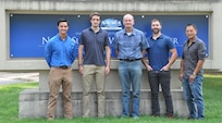 IMAGE: – Pre-emplaced Electric Vehicle Stopper (PEVS) Primary Engineer Kevin Arrington, PEVS Engineer Joshua Rawles, PEVS Project Lead David Dvorak, Booz Allen Hamilton Contract Engineer Kevin Komiensky and PEVS Technical Lead Dr. Yeong-Jer Chen stand in front of the NAVSEA Warfare Centers sign at Naval Surface Warfare Center Dahlgren Division.