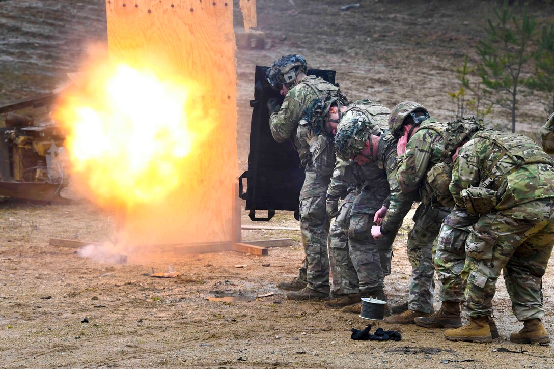 Soldiers stand huddled together in a line as an explosive detonates.