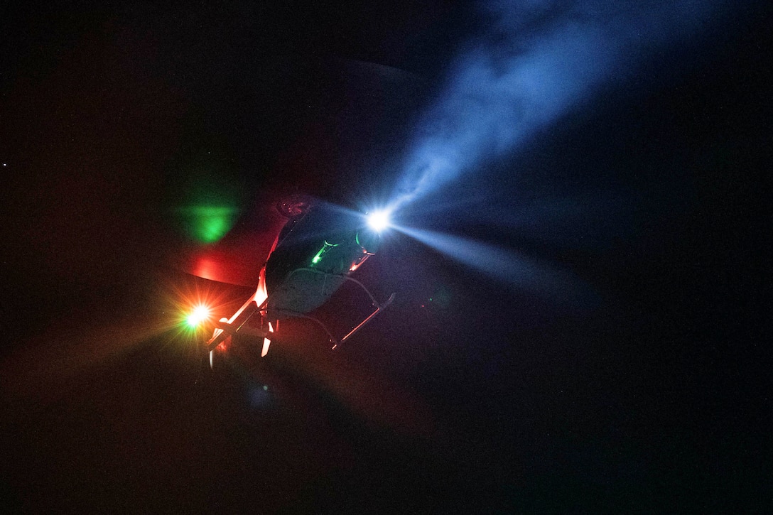 A helicopter hovers over a ship illuminated by colorful lights.