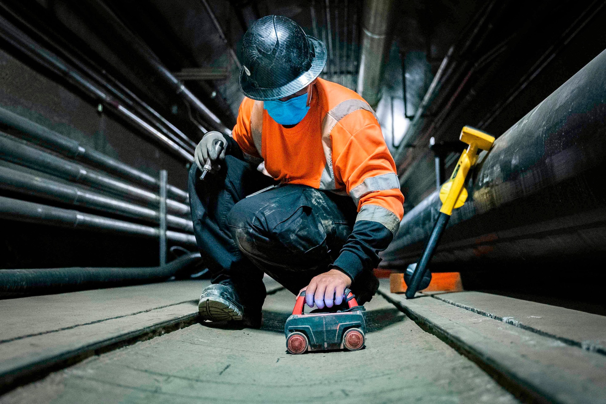A contractor wearing a face mask kneels down to use a tool.