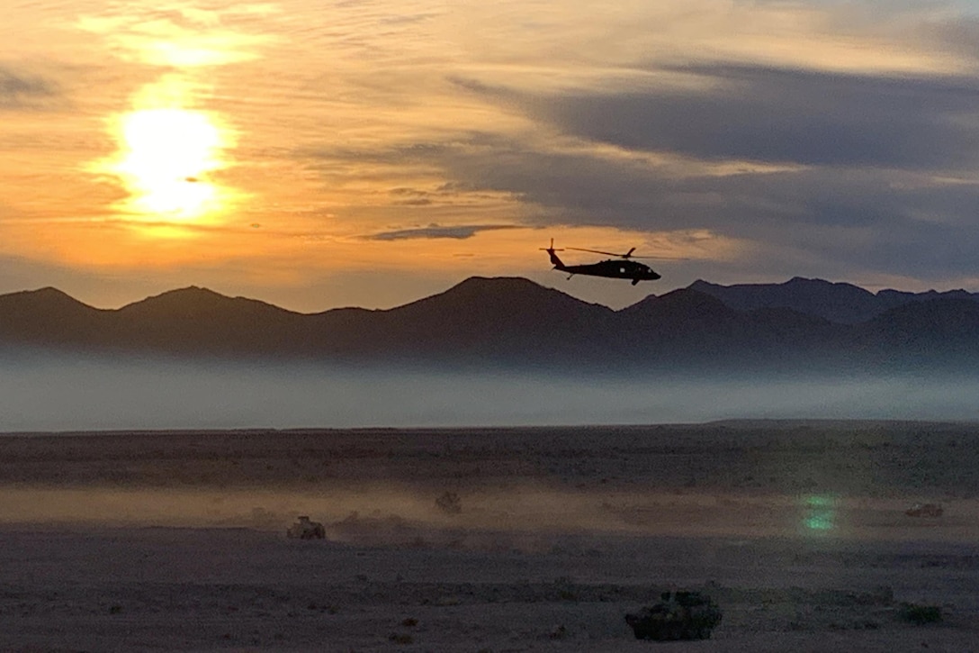 Tanks move across a desert and a helicopter flies across the sky at sunrise.