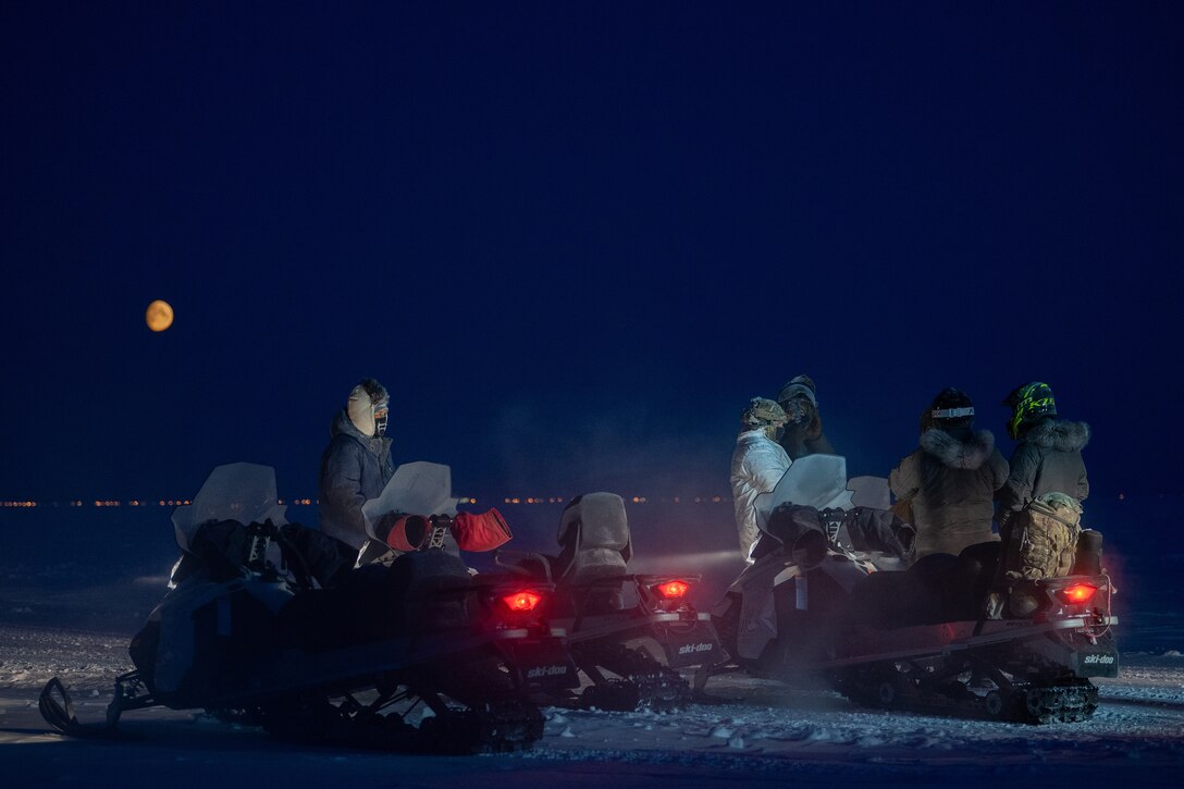 Airmen and snowmobiles are gathered on the ice during a moonlit night.