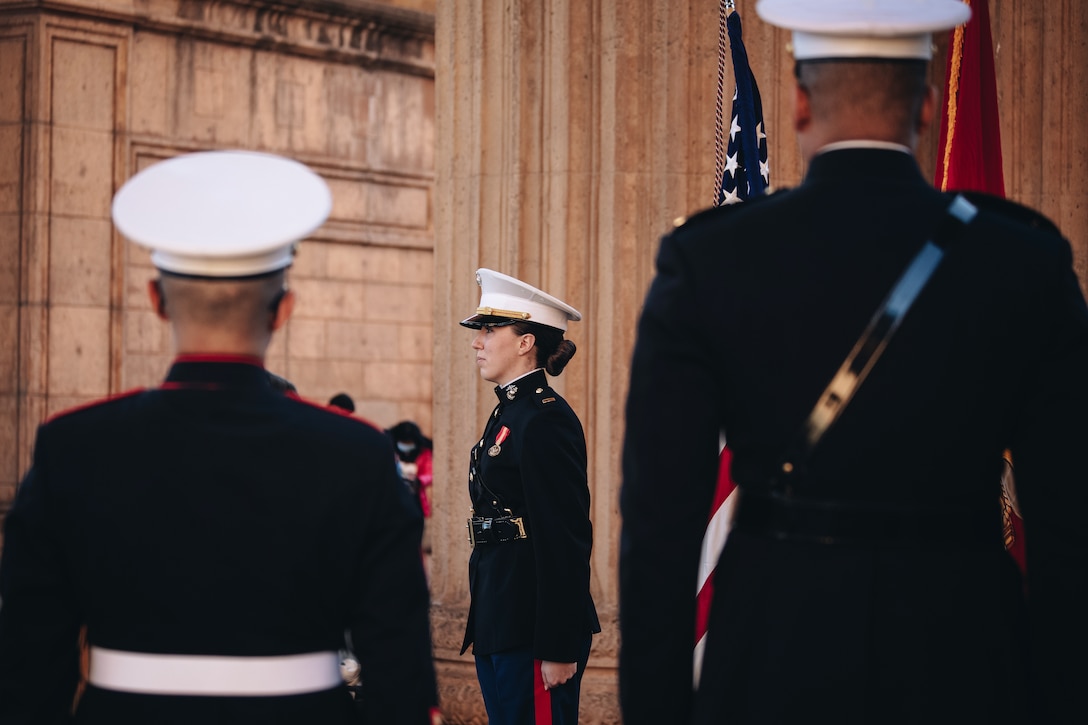 A Marine Corps officer stands at attention during a ceremony. Two other Marines stand with their backs to the camera.