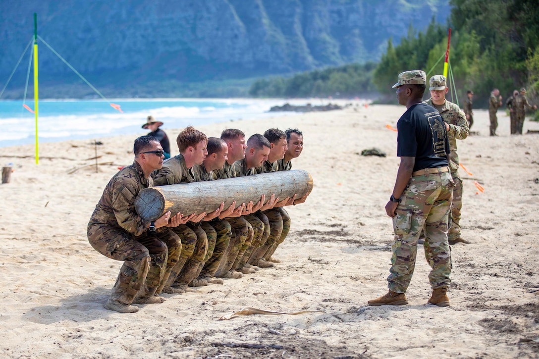 Soldiers do squats while holding a log as others watch.