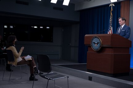 A man speaks at a news conference. A seated woman raises her hand slightly.