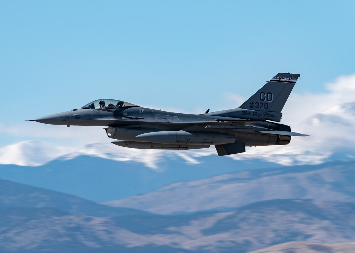 An F-16C Fighting Falcon flies against a backdrop partly cloudy skies and snow-capped mountains.