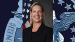 White woman with blond hair in a business suit with the US Flag behind her.