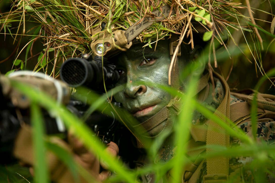 A Marine in camouflage aims a weapon in the woods.