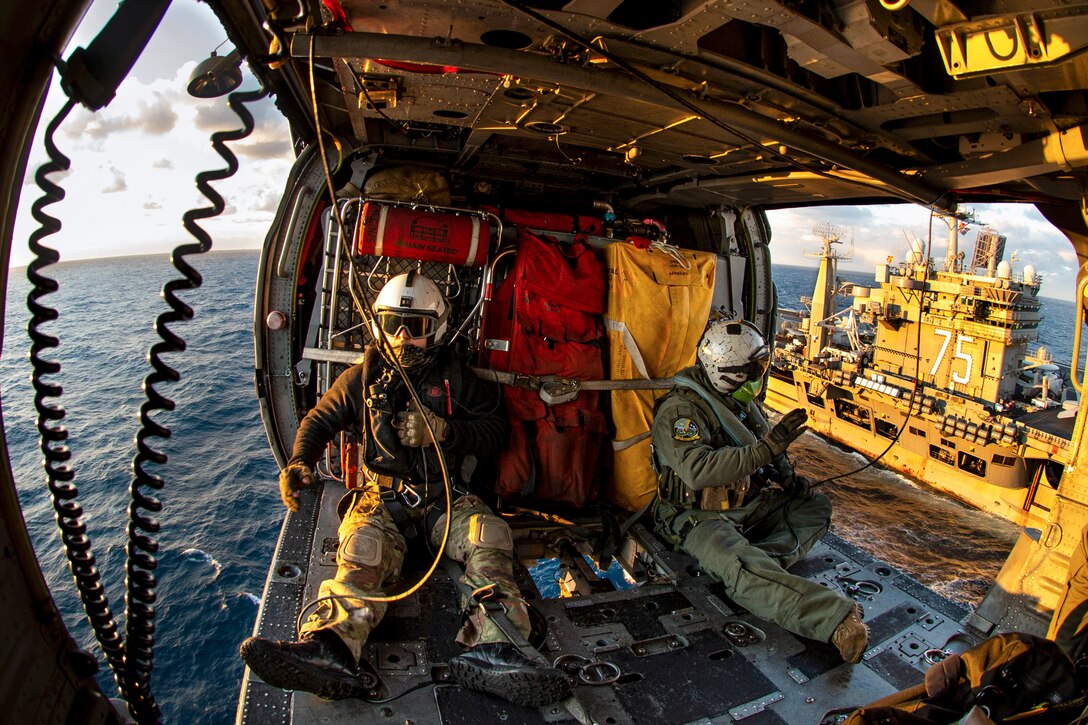 Two sailors sit in an airborne helicopter while flying next to a ship at sea.