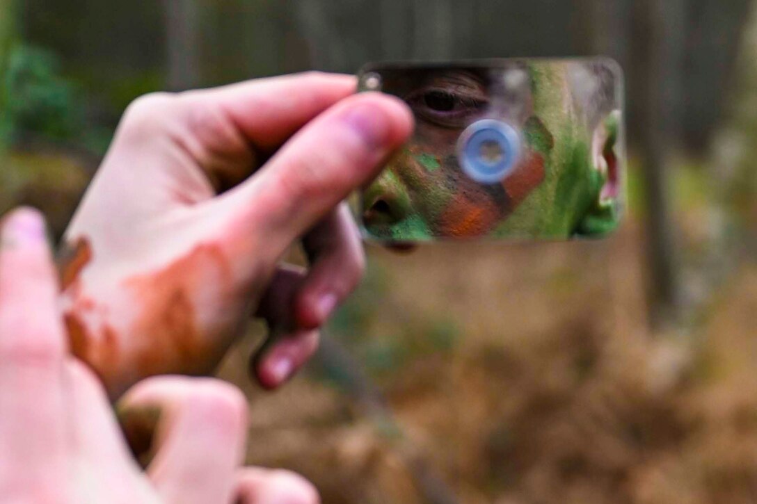 An airman uses a small mirror to apply camouflage face paint.