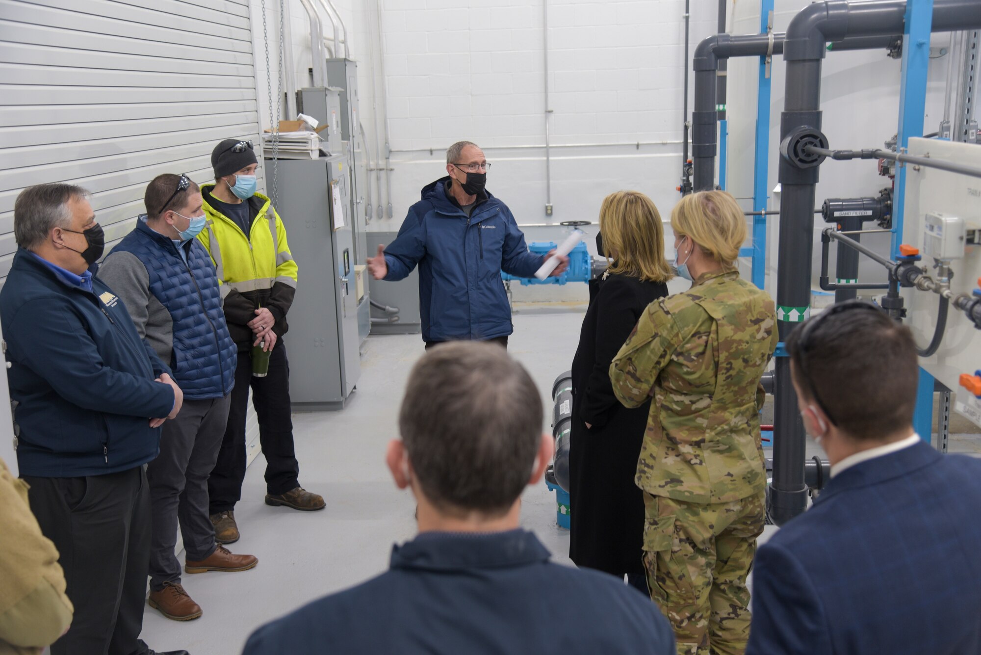 A man in a blue jacket briefs a group of people. One woman in the group has on an Air Force uniform.