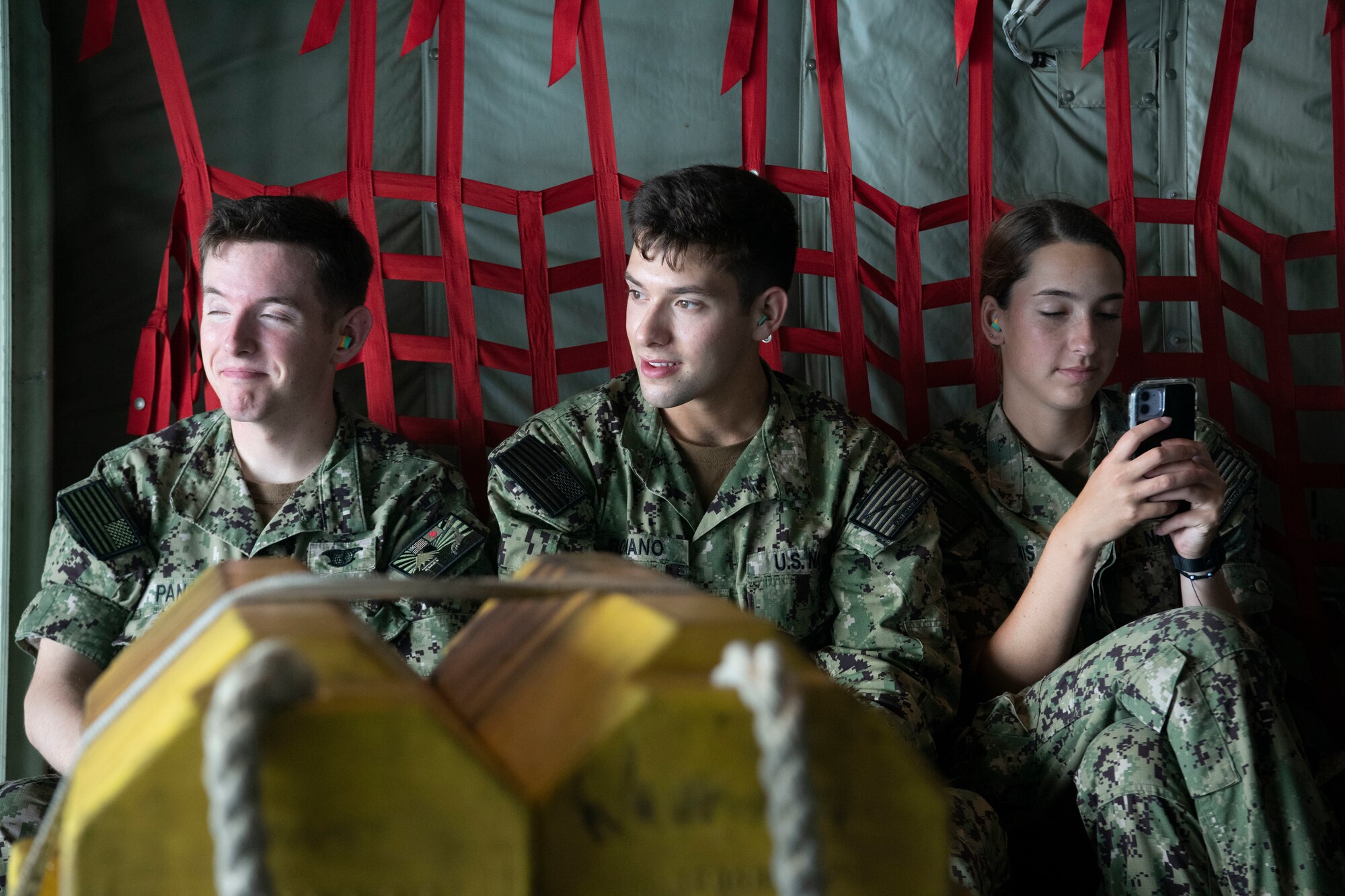 Three navy cadets sit in the cargo section of a military cargo plane.