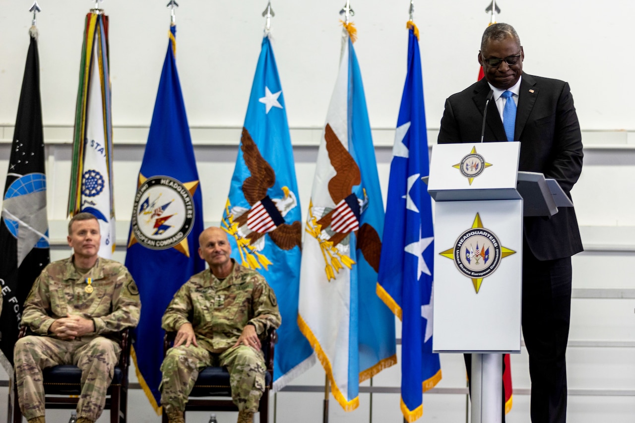 A man speaks from a stage while two other men in uniform sit in chairs behind him.