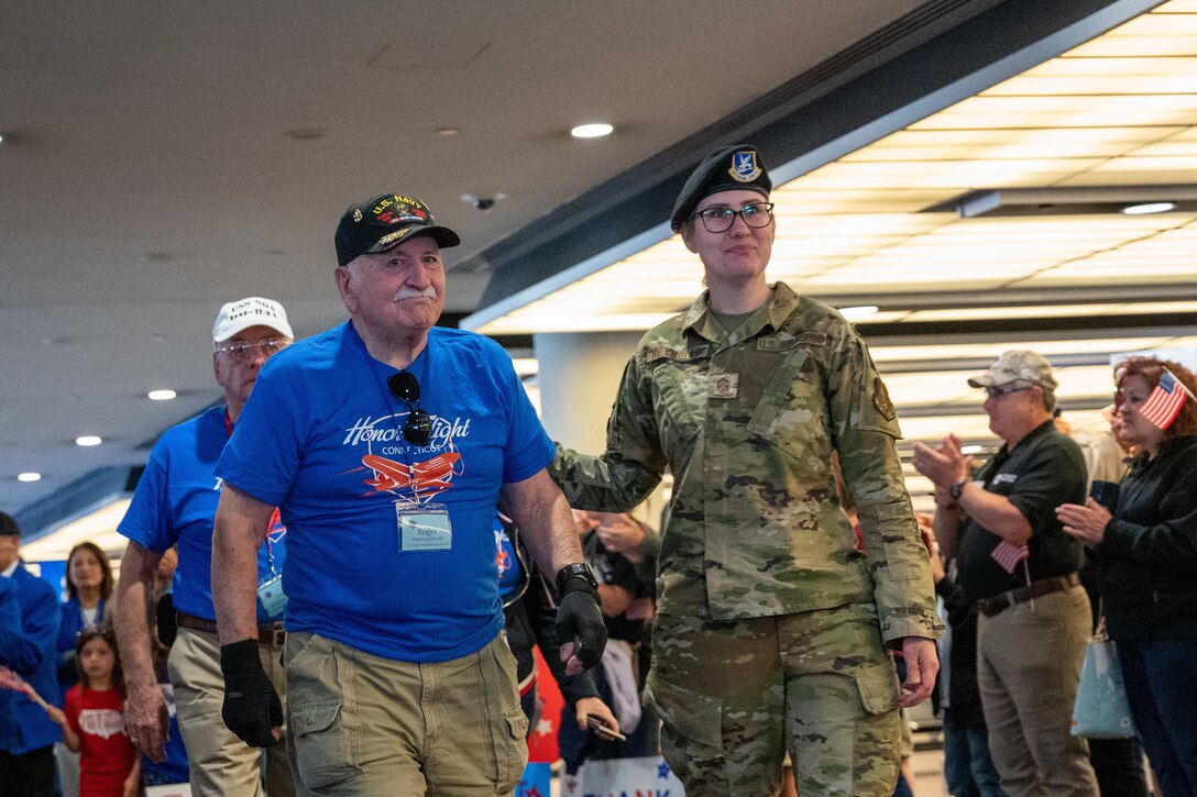 An airman escorts a man through an airport as people clap for them.