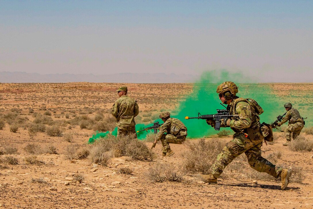 Soldiers move through desert terrain while green smoke rises behind them.