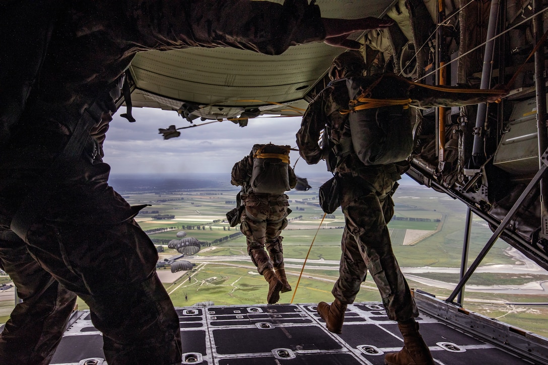 Soldiers with parachute packs walk toward the back of an open aircraft. Actively parachuting service members can be seen in the background.