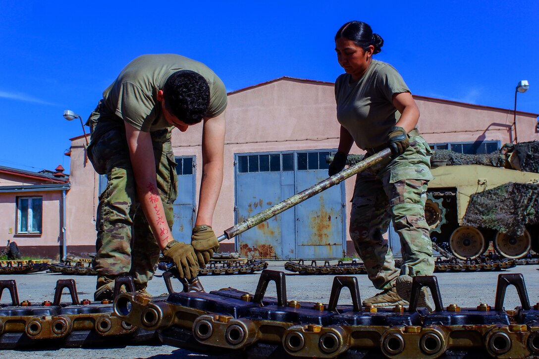 Two soldiers work together to use a tool on tank tracks.