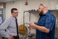 Ryan Franke (right), a technician in Carderock’s Subtractive and Additive Manufacturing Branch, explains to Steve Ouimette, Carderock’s Deputy Technical Director, what Benchy the Tug Boat is and what it is used for, during the grand opening of Carderock’s new Advanced Manufacturing Prototyping (AMP) Lab. Benchy, or Benchmark, is the 3D printed part that Franke is holding. Benchy is a quick, little test part that was created to test the limits of a 3D printer. It was designed with a few features that are known to be difficult obstacles for 3D printers. Here, Franke explains that Benchy is used in the AMP Lab training class to allow new users the opportunity to get in the Driver’s Seat and learn start to finish how to create a Build Platform for the Printers, and how to get that platform from the computer to the printer itself and how to get the print started. (U.S. Navy photo by Devin Pisner)