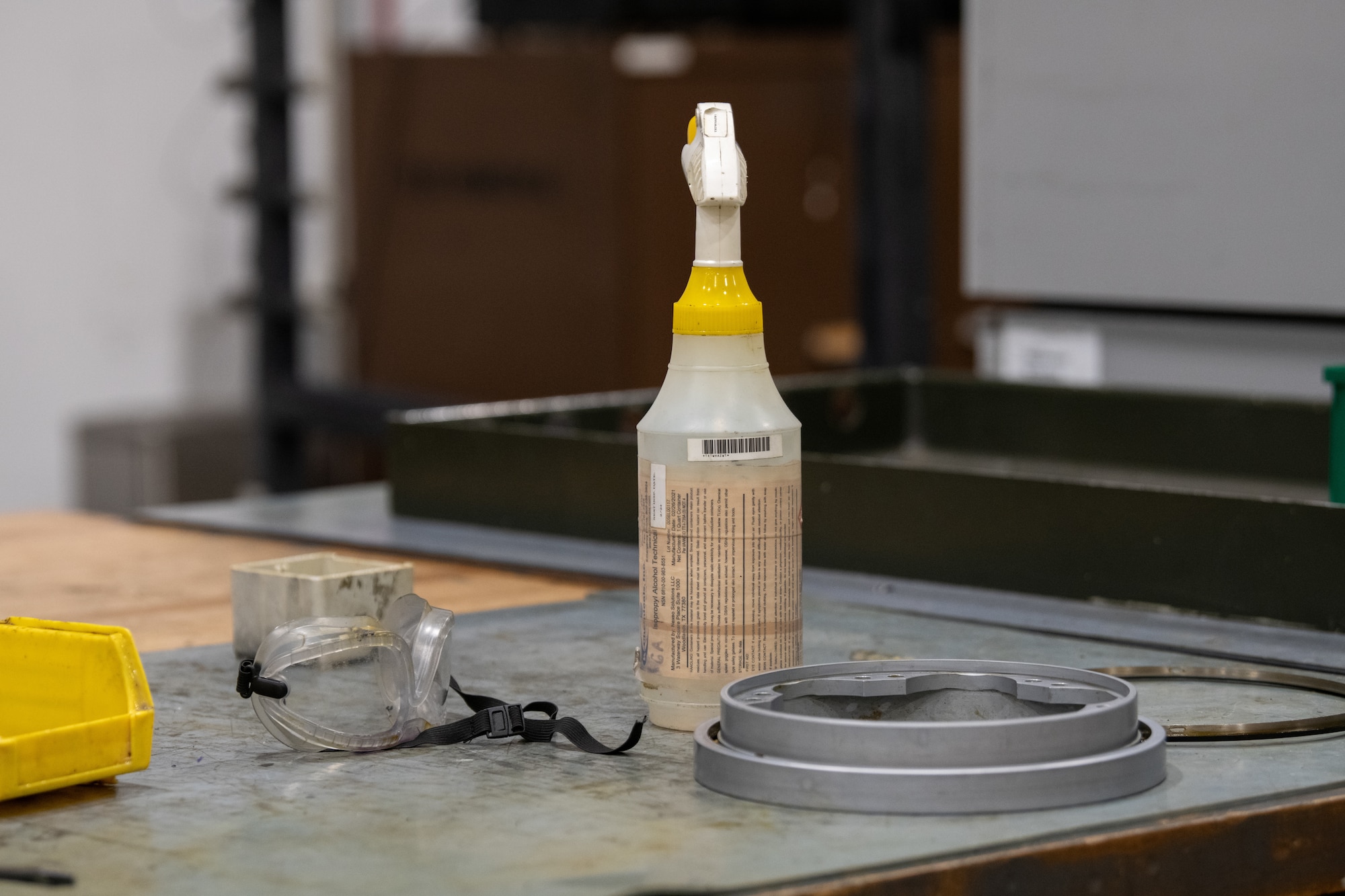Cleaning spray, safety equipment, and weapon system part sit on table