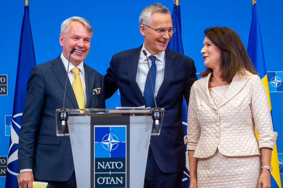 Three smiling civilians stand behind a lectern with a sign that says "NATO."