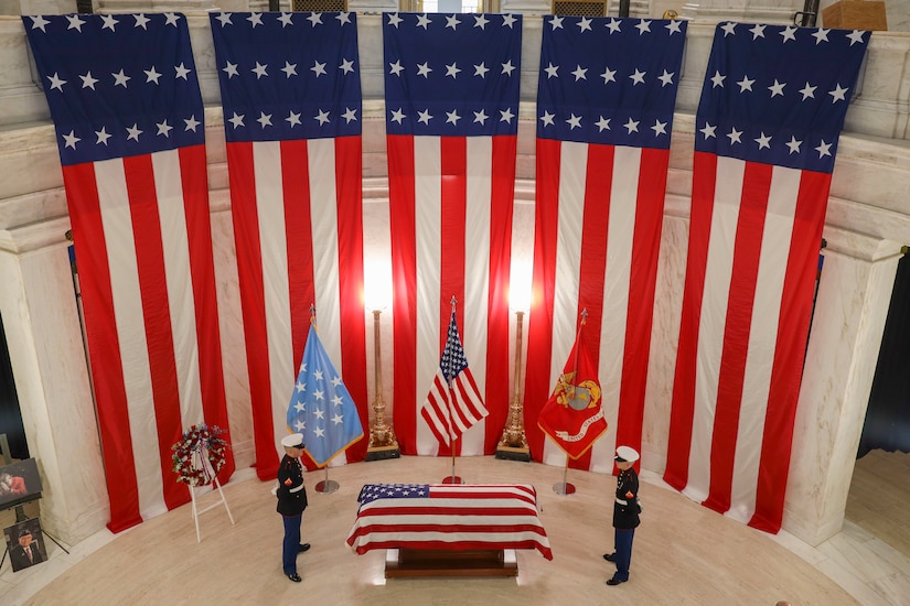 Two Marines stand on either side of an American-flag-draped coffin and a round room.