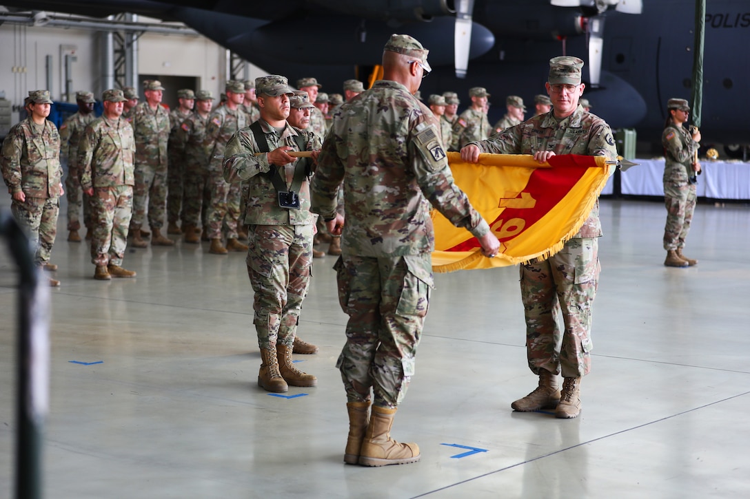 Two soldiers furl a yellow and red flag while a formation of soldiers stands at attention in the background.