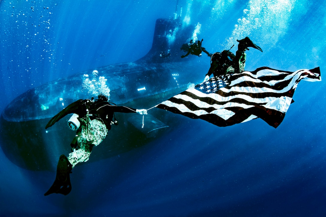 Navy divers swim with an American flag by a submarine.