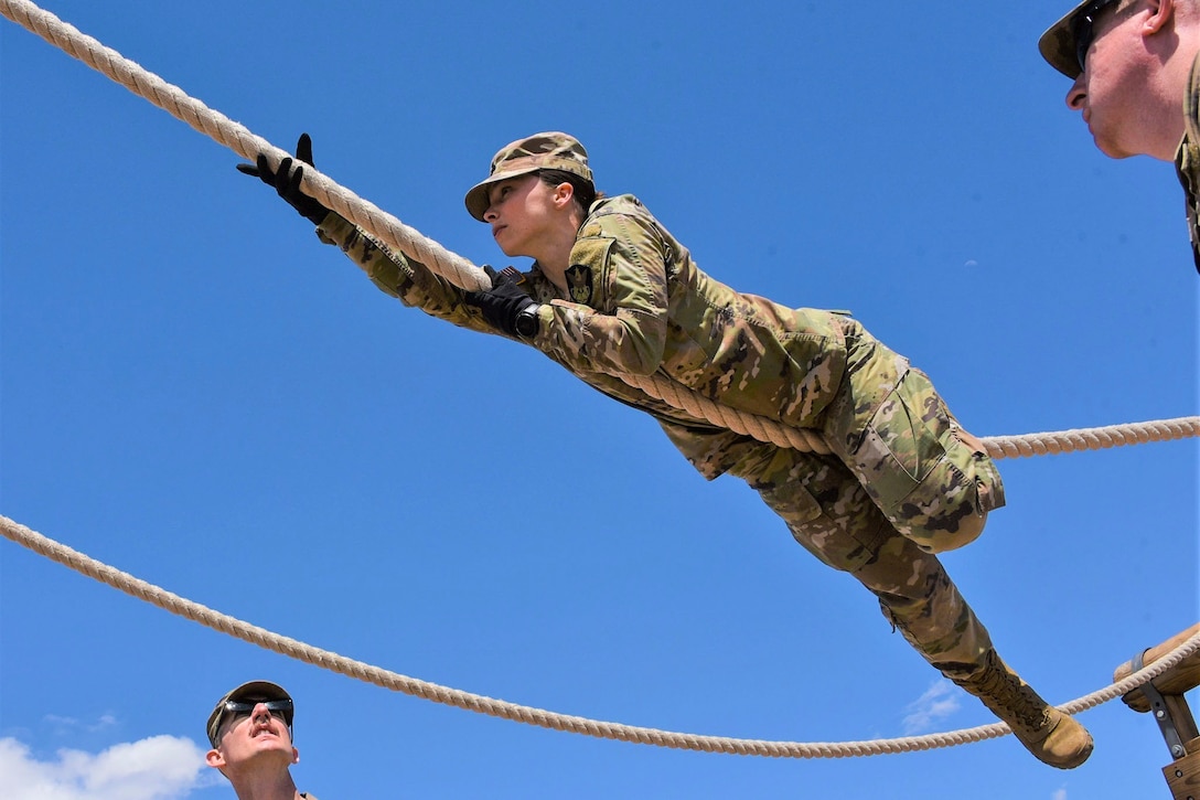 A soldier moves across a rope obstacle suspended in air as two others watch.