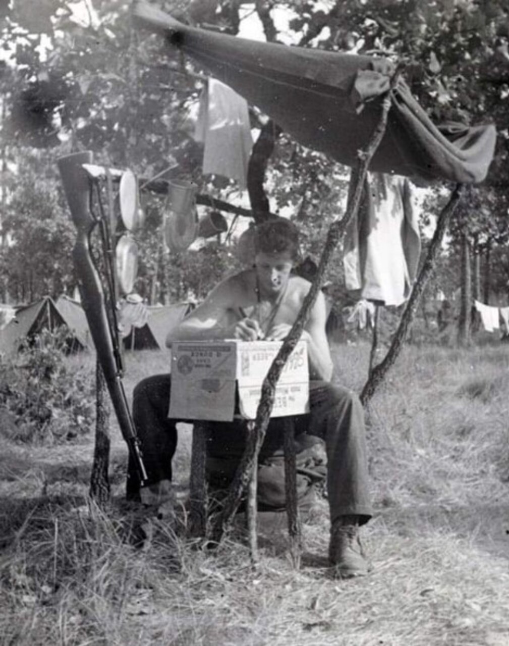 A man writes a letter on a box while under a makeshift awning.