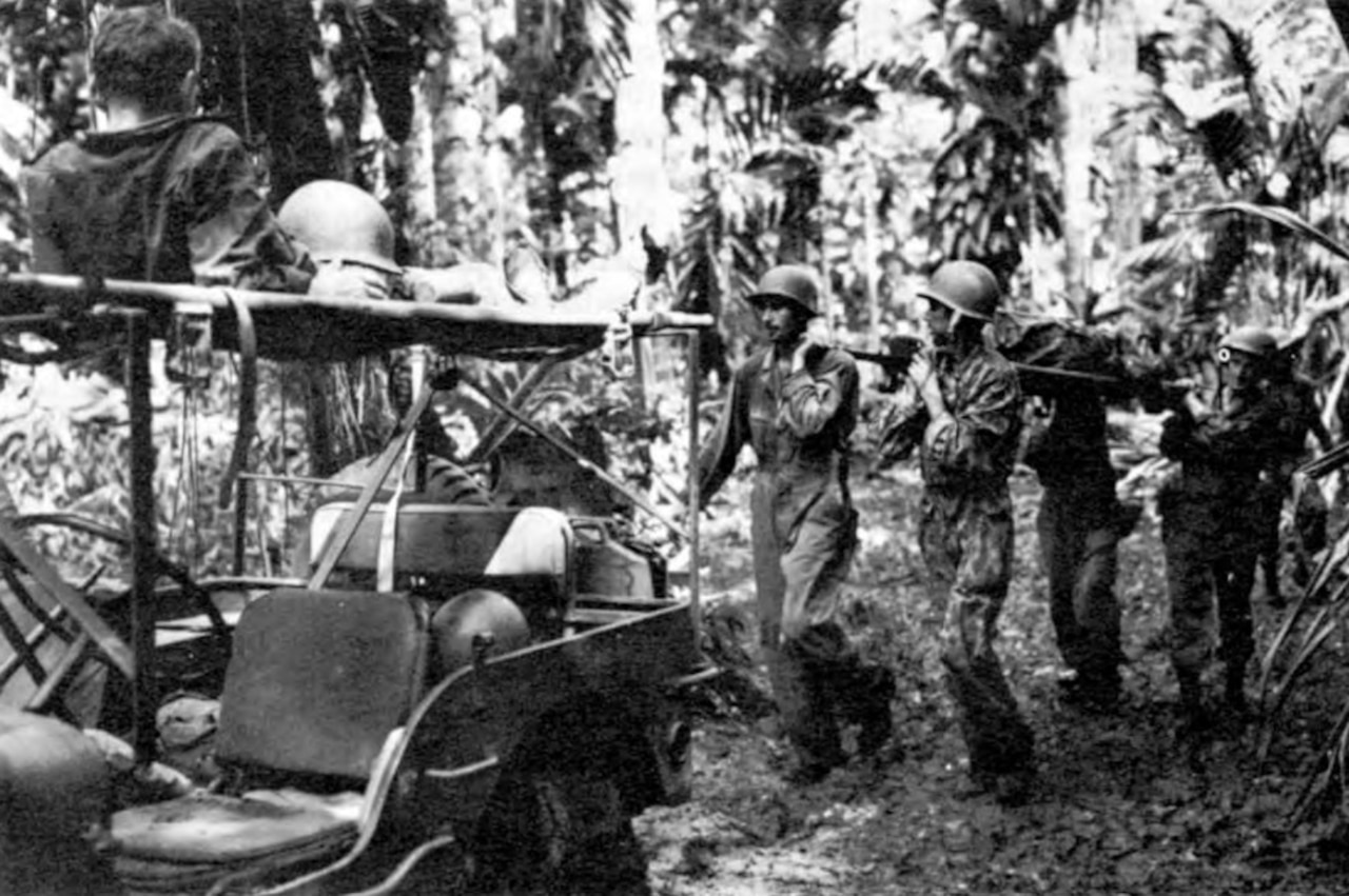 Four men carry a person on a stretcher through jungle toward a Jeep on which another man is laying.