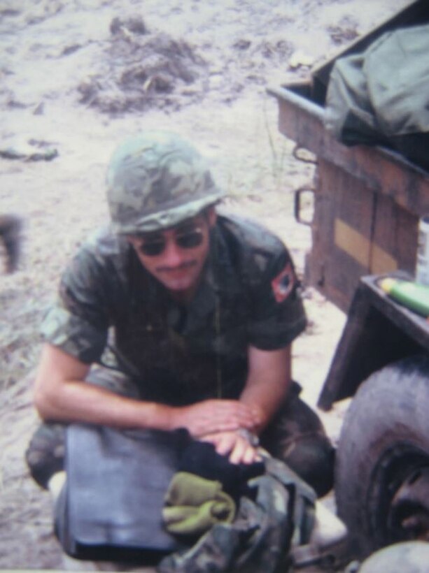 A soldier wearing sunglasses and helmet crouches on ground.