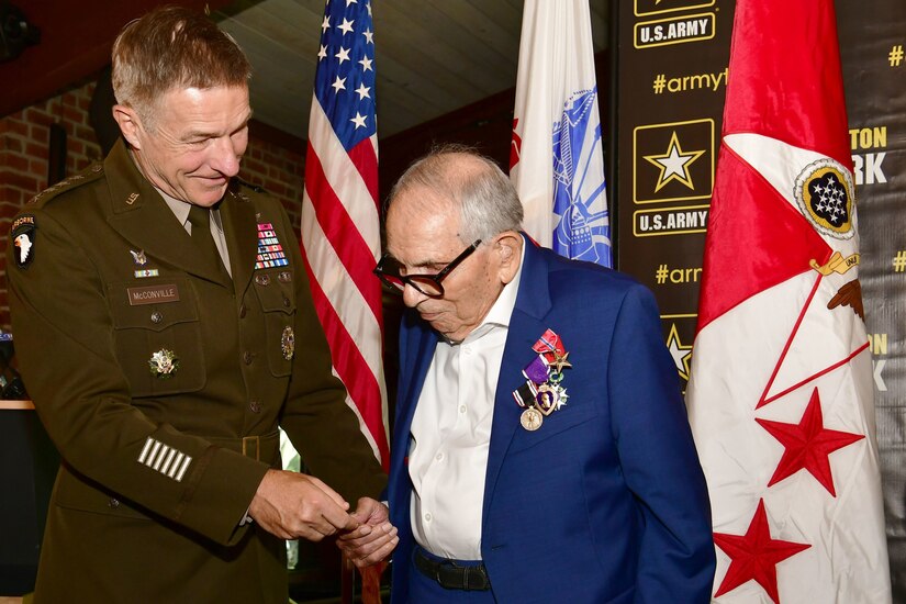 A man in uniform and an older man stand in front of some flags.