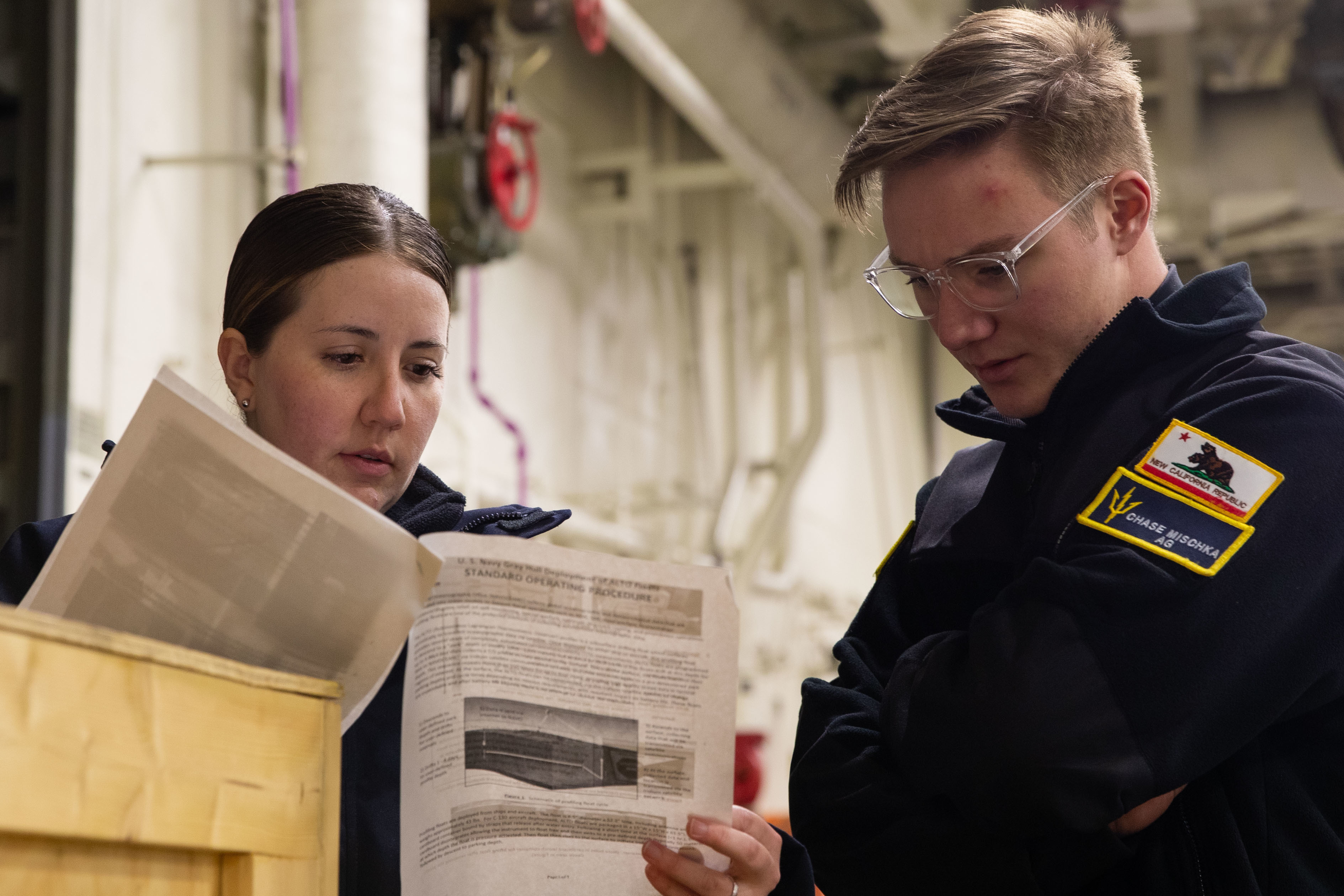 220514-N-CM110-1006 PACIFIC OCEAN (May 14, 2022) – Aerographer’s Mate 1st Class Ashley Mccown, from El Paso, Texas, left, and Aerographer’s Mate 3rd Class Chase Mischka, from Santee, California, read instructions for unboxing a buoy in the vehicle stowage area aboard amphibious assault carrier USS Tripoli (LHA 7), May 14, 2022. Tripoli is underway conducting routine operations in U.S. 7th Fleet. (U.S. Navy photo by Mass Communication Specialist 3rd Class Maci Sternod)