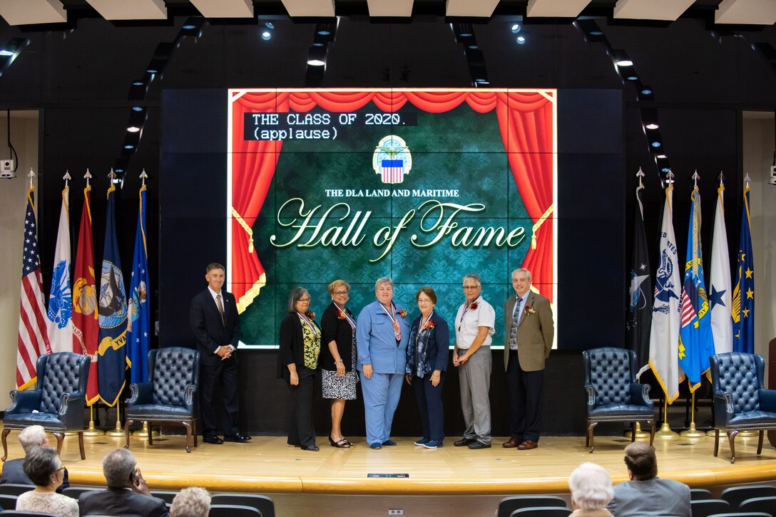 Deputy Commander watson stands with five women and a man in business dress on a stage. The man does not wear a medal. The five women are wearing medals.