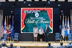 Two women and a man in business dress on a large stage wearing medals.