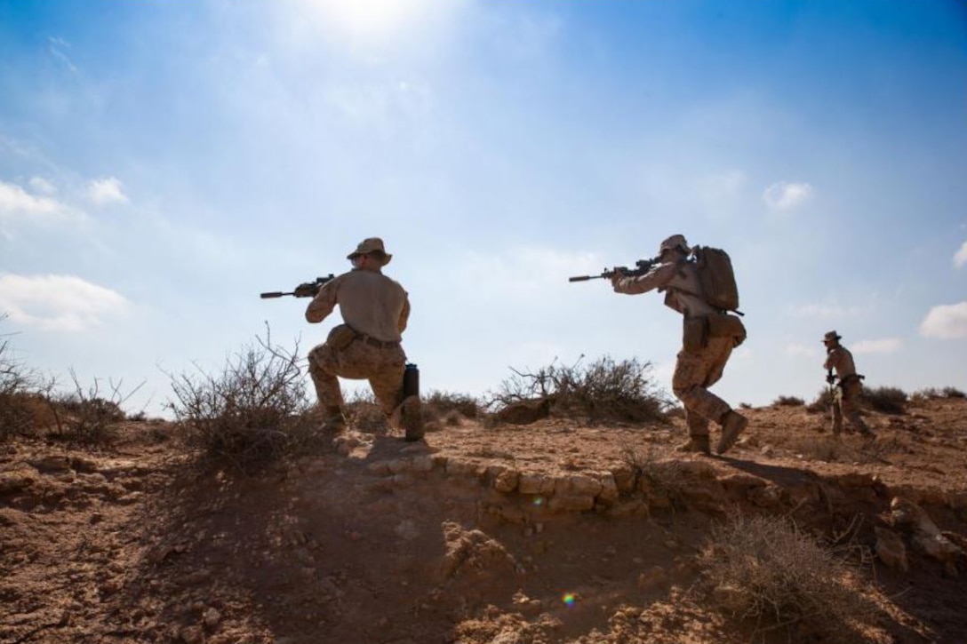 Marines holding weapons walk cautiously across a dirt terrain.