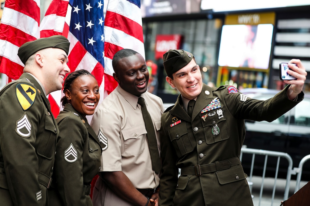 Four soldiers pose for a selfie in front of American flags on a city block.