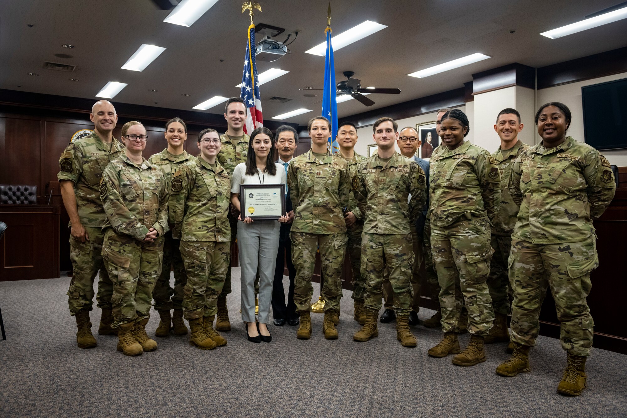 Group of military members in uniform alongside civilians hold up a certificate in a courtroom.