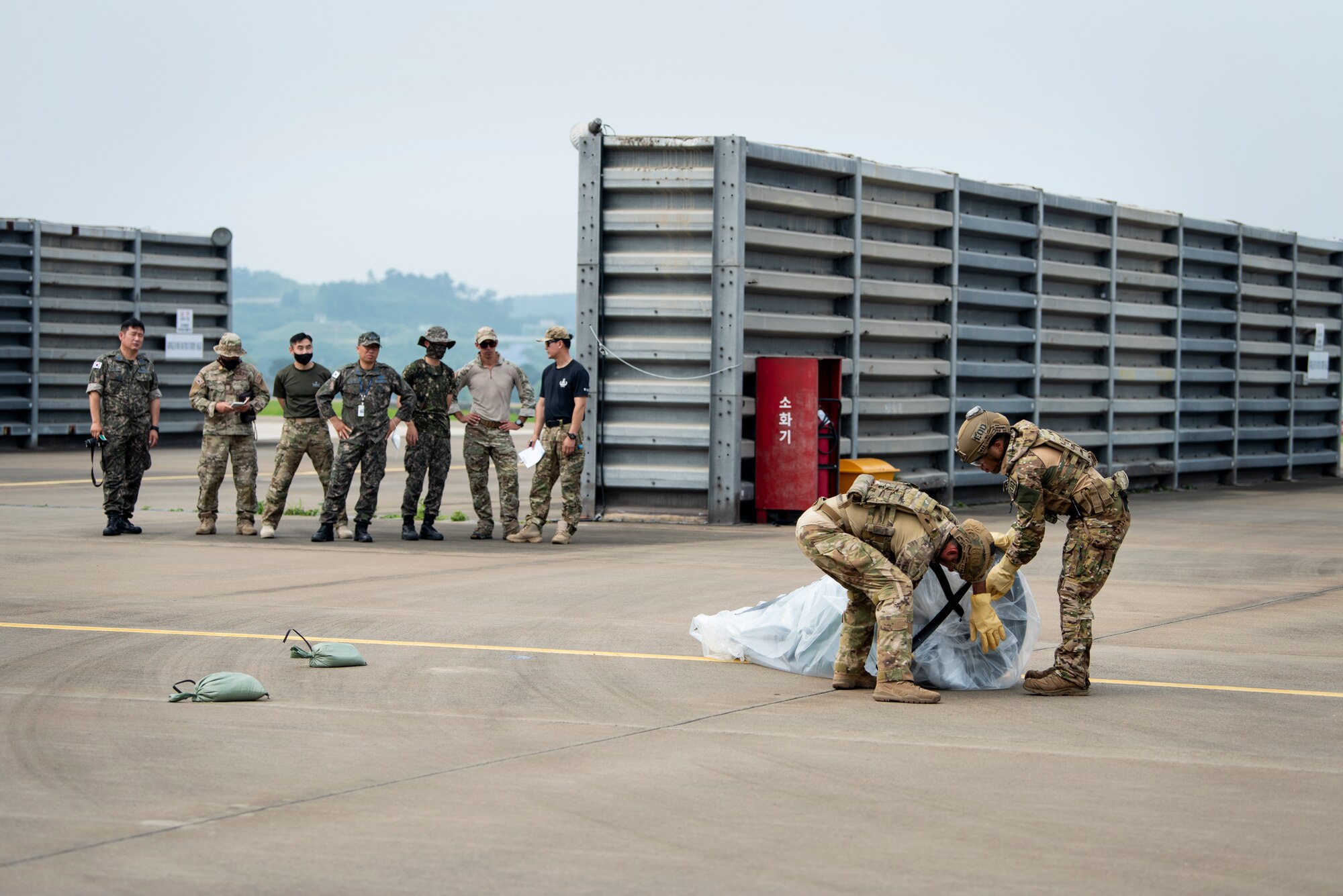 Members of the Republic of Korea Air Force, Explosive Ordnance Disposal team, observe as U.S. Air Force Staff Sgt. Michael Augustus and Senior Airman Joshua Young, 35th Civil Engineer Squadron, Explosive Ordnance Disposal technicians from Misawa Air Base, Japan, practice covering a mock chemical weapon during a bilateral training event at Suwon Air Base, Republic of Korea, July 6, 2022.
