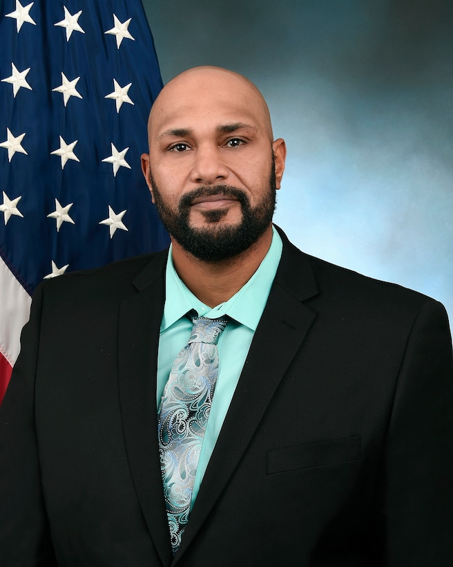 Official photo of a man in a black suit jacket and teal shirt in front of an American flag and blue background.