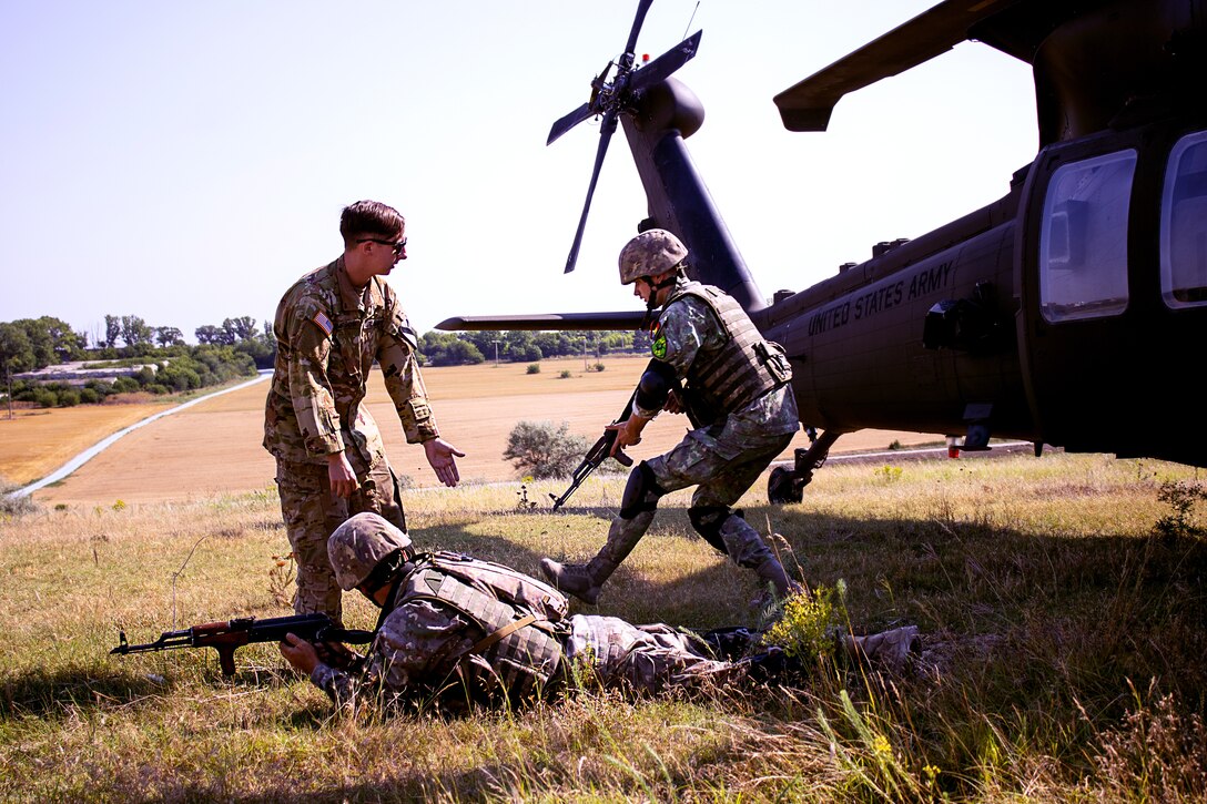 A  service member gives instruction to two foreign uniformed service members in a grassy area with a helicopter in the background.